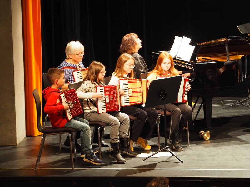 Eine Gruppe von Personen, darunter Kinder und Erwachsene, sitzt auf der Bühne im großen Saal des Uditoriums und spielt auf Instrumenten, die sie in der Musikschule der Stadt Uhingen gelernt haben.