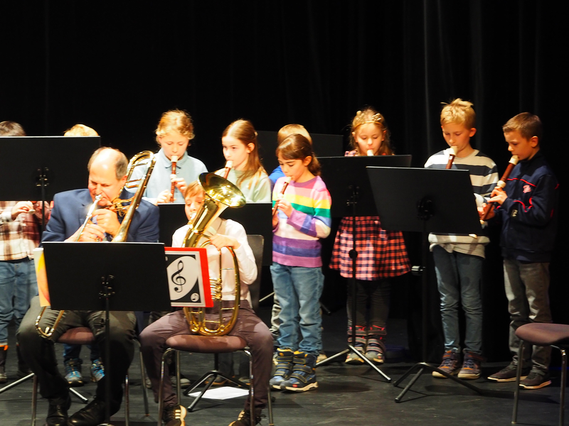 Eine Gruppe von Kindern und ein Erwachsener spielen verschiedene Blasinstrumente gemeinsam auf der Bühne im großen Saal des Uditoriums beim Adventskonzert der Musikschule.