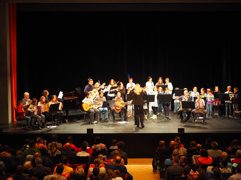 Eine Gruppe von Musikern, darunter Kinder, Jugendliche und Erwachsene, spielt auf der Bühne des Uditoriums vor einem Publikum verschiedene Instrumente beim Adventskonzert der Musikschule. 