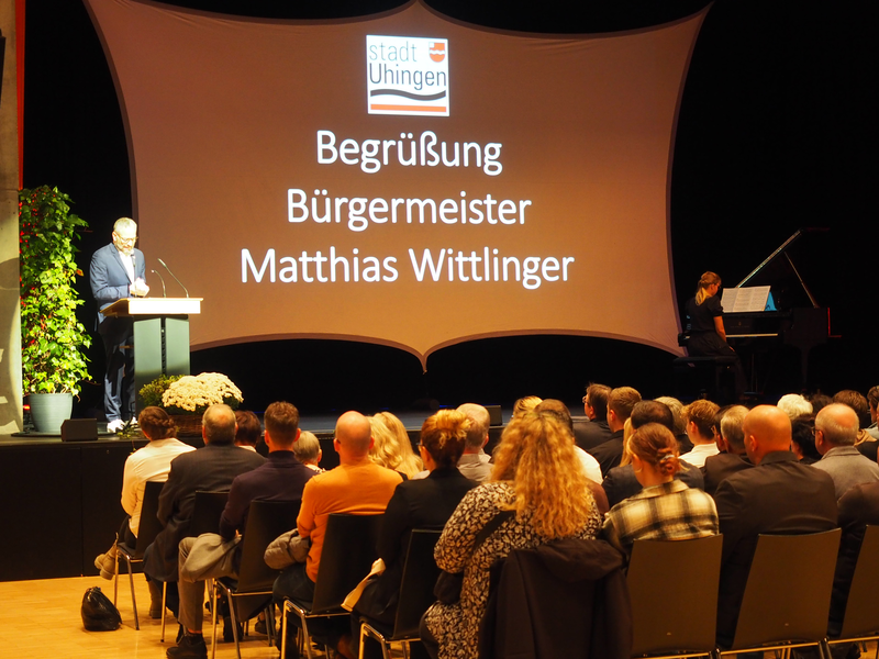 Auf der Bühne eines Festsaals hält Bürgermeister Matthias Wittlinger am Rednerpult eine Rede, während im Hintergrund eine Leinwand mit der Aufschrift "Begrüßung Bürgermeister Matthias Wittlinger" zu sehen ist, und links eine Frau Klavier spielt.