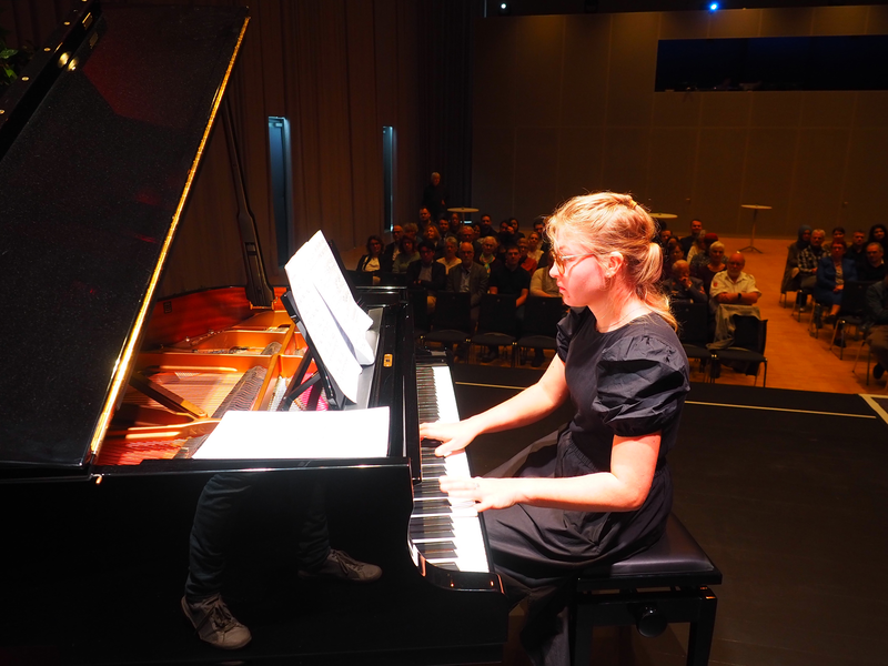 Eine Frau, Lidiia Baryshevska, in einem schwarzen Kleid spielt Klavier auf einer Bühne vor einem sitzenden Publikum.