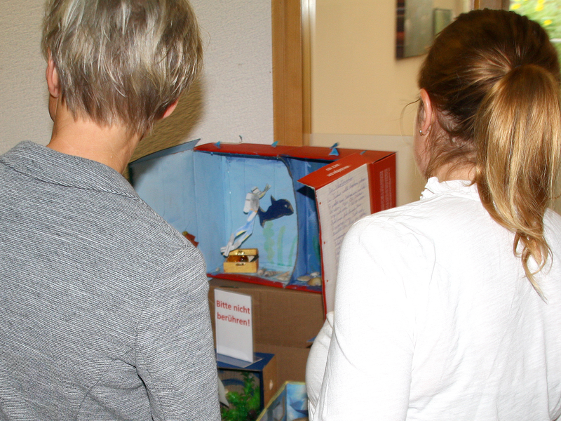 Zwei Frauen, Mitglieder der Jury, schauen sich ein Diorama an. In einem Karton hat ein Kind eine Unterwasserlandschaft gebastelt.