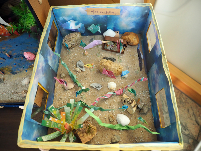 Aus Papier gefaltete Fische und Seeschlagen, sind auf einem sandigen Boden in einem Diorama. In einer Schatzkiste funkeln Schätze.