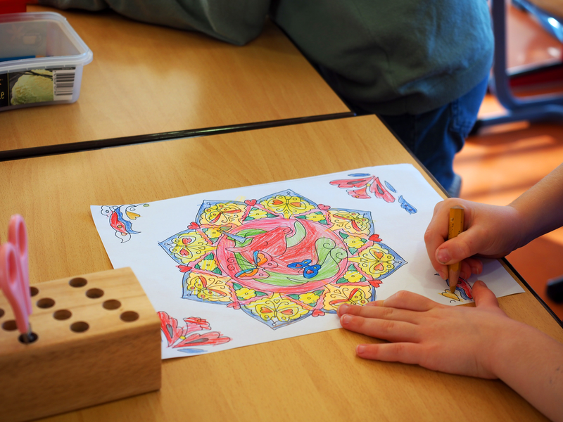 Ein Kind malt ein buntes Mandala auf einem Blatt Papier, das auf einem Tisch liegt.