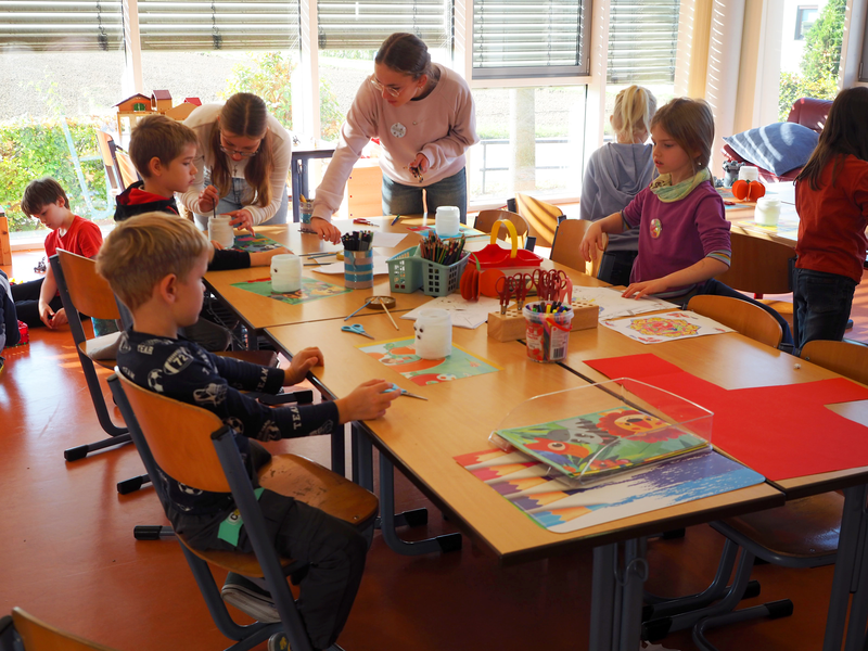 In einem sonnigen Klassenraum sitzen in der Ferienbetreuung in der Grundschule Sparwiesen mehrere Kinder an einem Tisch und basteln mit verschiedenen Materialien, während eine erwachsene Person den Kindern hilft.