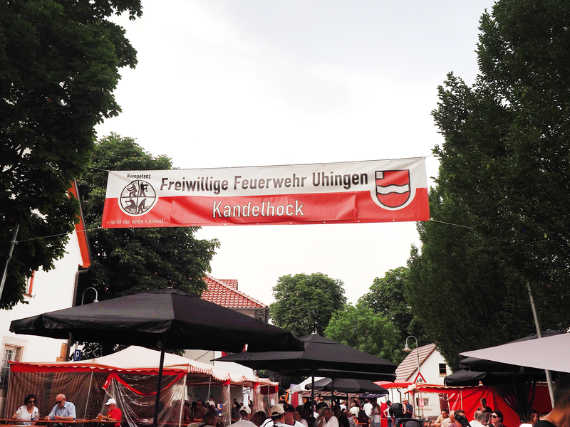 Unter einem zwischen Bäumen gespannten Banner mit der Aufschrift "Freiwillige Feuerwehr Uhingen Kandelhock" stehen Biertische und Bierbänke, auf denen Menschen sitzen.