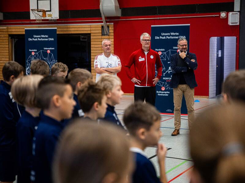 Andreas Henning und Thomas Escher von der Schüssler Escher Stiftung stehen in einer Sporthalle neben Uhingens Bürgermeister Matthias Wittlinger. Vor ihnen stehen Kinder und Jugendliche.