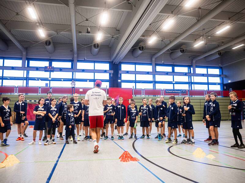 In einer Turnhalle stehen mehr als 30 Kinder und Jugendliche in Sportkleidung vor einem Sportler, der ihnen eine Aufgabe erklärt.