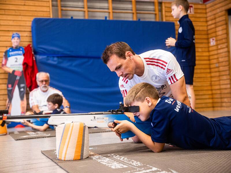 Ein Junge liegt in einer Halle auf einer Matte und schaut durch das Zielrohr eines Gewehrs. Neben ihm kniet Simon Schempp und gibt dem Jungen Tipps.