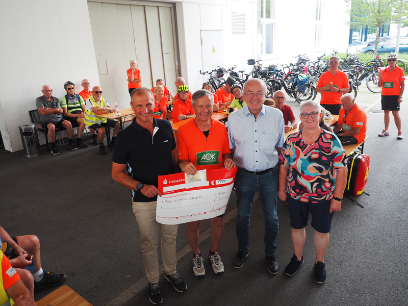 Vier Männer halten einen Spendenscheck, hinter ihnen sitzen an Bierbänken Radfahrer.