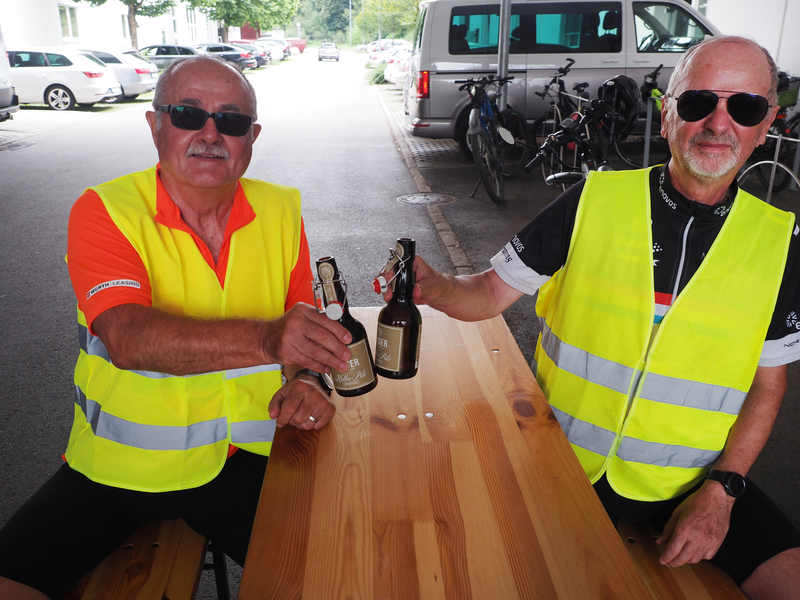 Zwei Männer sitzen nach einer Radtour an einem Biertisch und stoßen mit Bierflaschen an.