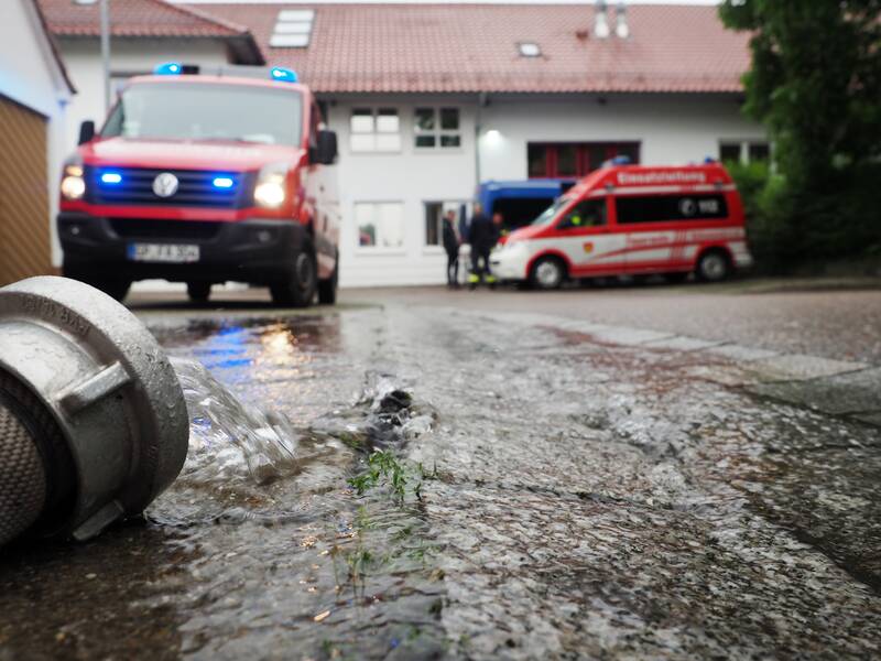 Aus einem Feuerwehrschlauch läuft Wasser auf die Straße, im Hintergrund stehen Einsatzfahrzeuge der Feuerwehr.