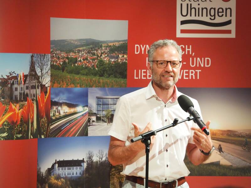 Bürgermeister Matthias Wittlinger spricht in ein Mikrofon und steht vor einer roten Wand, auf der Impressionen von Uhingen zu sehen sind.