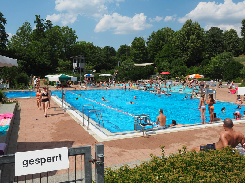 Im Schwimmerbecken des Uhinger Freibads schimmert das Wasser azurblau und viele Menschen halten sich auf. Sie schwimmen oder hüpfen hinein. Im Hintergrund sind Büsche und Bäume.