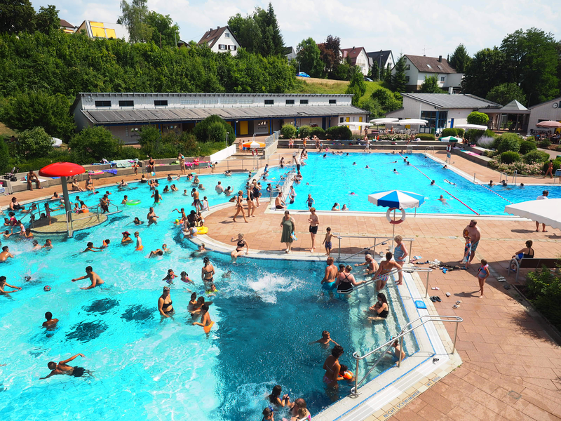 Im Nichtschwimmerbecken des Uhinger Freibads schimmert das Wasser azurblau und viele Menschen halten sich auf, spielen Ball, hüpfen hinein. Im Hintergrund sind Büsche, das Freibadgebäude und Bäume sowie Häuser.