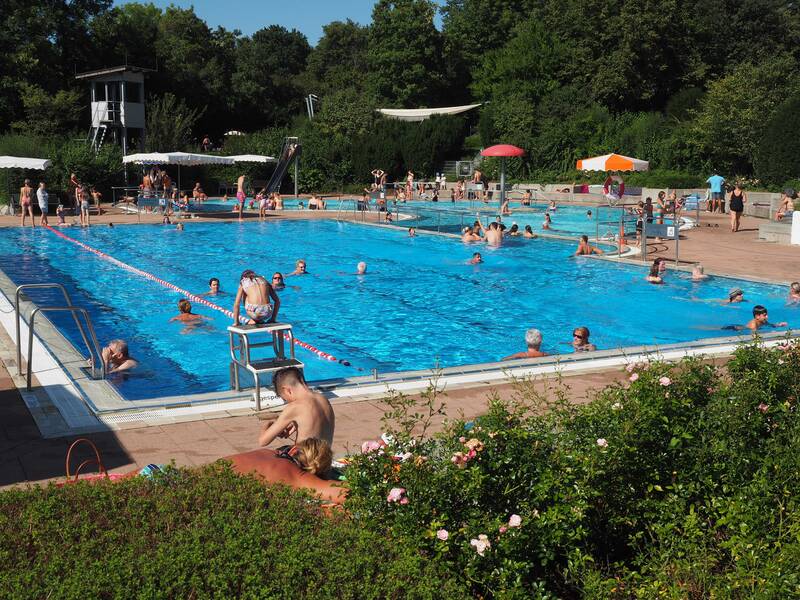 Im Schwimmerbecken des Uhinger Freibads schimmert das Wasser azurblau und viele Menschen halten sich auf. Sie schwimmen oder hüpfen hinein. Im Hintergrund sind Büsche und Bäume.