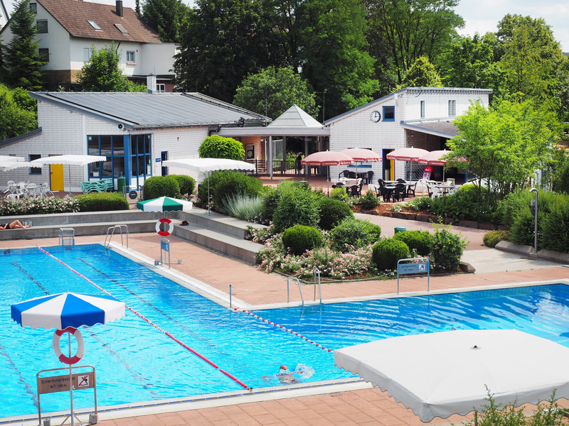 Das Freibadgelände mit dem Schwimmbecken und Sprungbecken, in denen Wasser ist. Büsche und Sträucher an gepflasterten Wegen und Betonstufen zum Liegen oder sitzen trennen den Badebereich vom Kiosk, vor dem Stühle und Tische unter Sonnenschirmen zum Verweilen einladen.