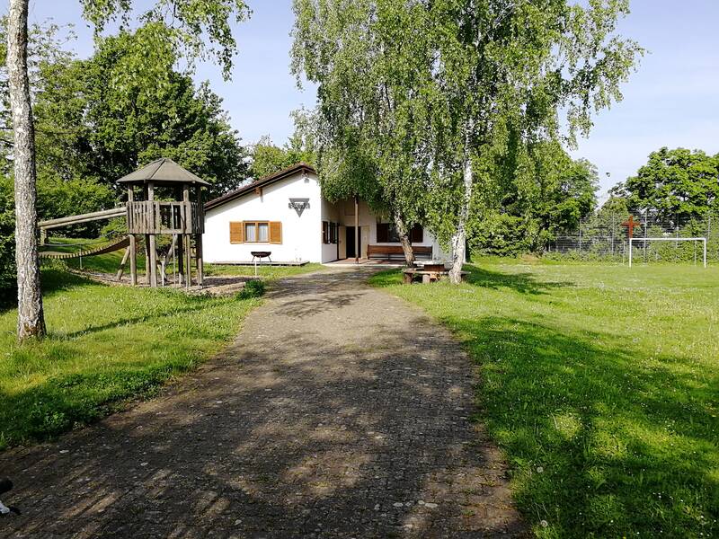 Giebelansicht vom weißes CVJM-Vereinsheim in Uhingen, davor eine Grillschale, links ein Spielturm mit Hängebrücke, rechts im Bild der Sportplatz mit Tor und metallenem Kreuz am Fangzaun